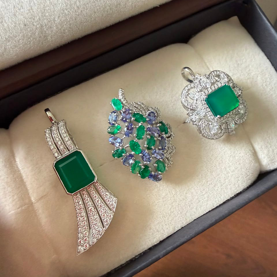 Three jewelry pieces displayed in a jewelry box: a silver pendant with a green rectangular stone, a silver and green leaf-shaped brooch with purple and green stones, and a silver ring with a green rectangular stone and clear accents.