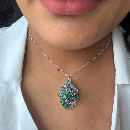 A close-up of a person wearing a silver chain necklace with a leaf-shaped pendant adorned with green, purple, and clear rhinestones, styled with a white collared shirt.