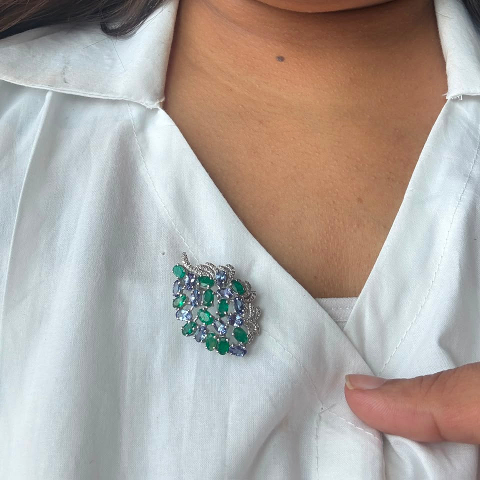 A close-up of a person wearing a silver-toned brooch with green and clear stones on a white collared shirt.