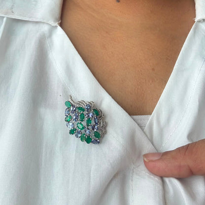A close-up of a person wearing a white collared shirt, featuring a heart-shaped brooch with green and blue gemstones set in silver on the chest.