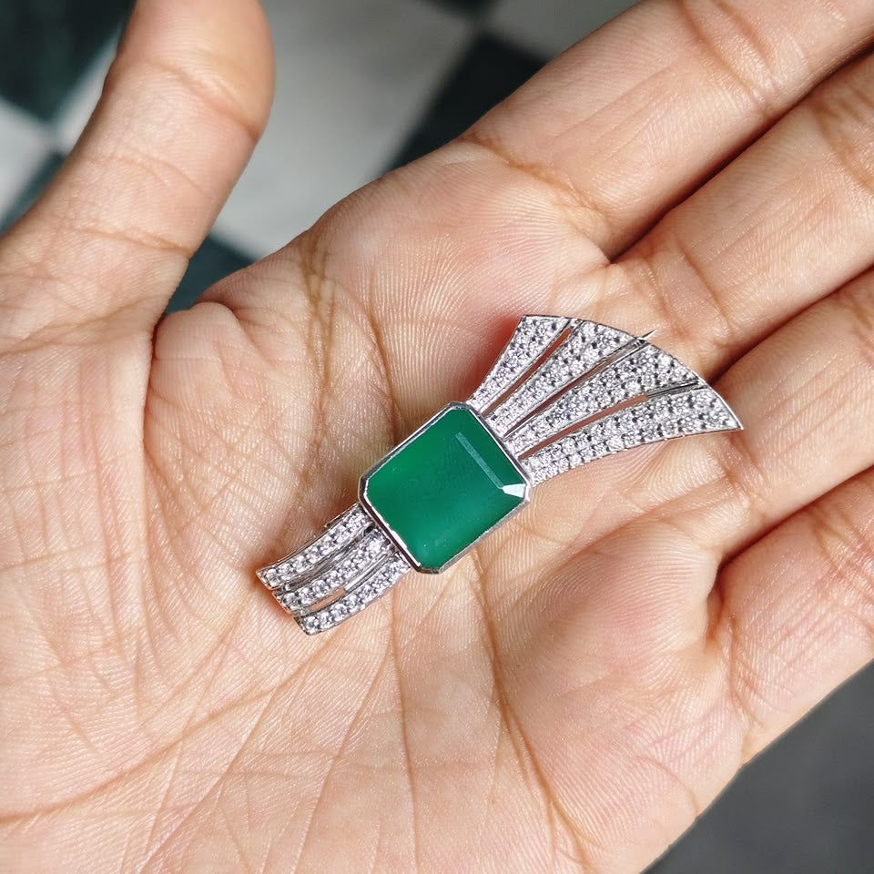 A silver-toned brooch with a rectangular green stone and a fan-like arrangement of small clear stones, displayed on an open hand.