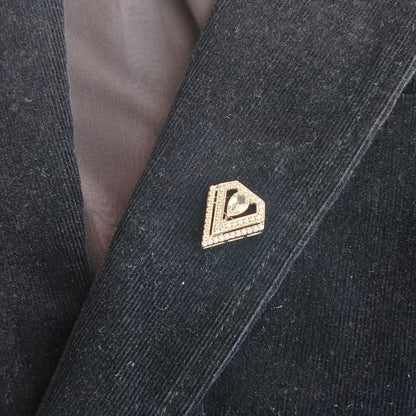 A close-up of a dark charcoal gray corduroy blazer jacket lapel with a small gold geometric pin featuring a central clear gemstone, fastened over a brown shirt.