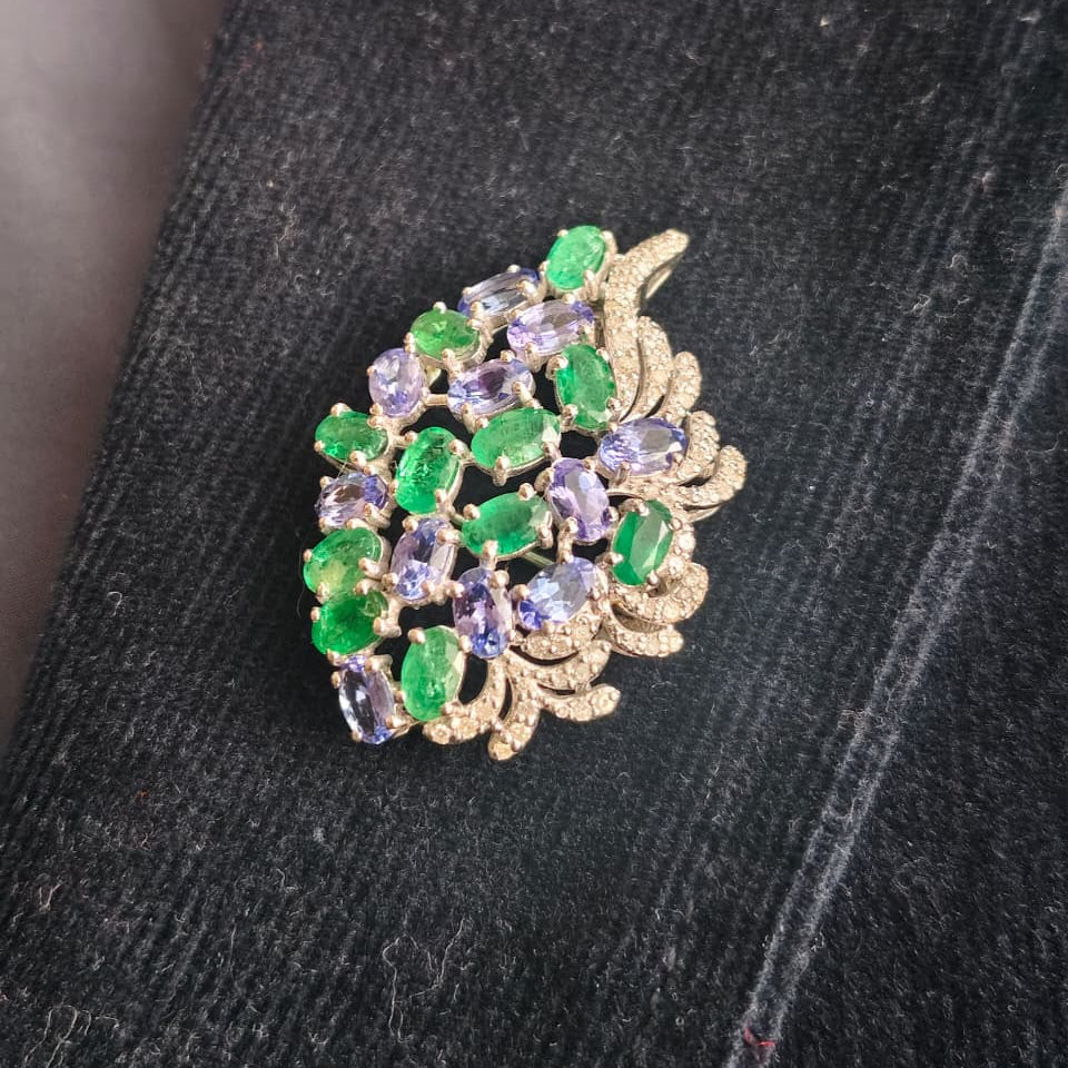 A vintage-style brooch featuring green and lavender purple crystals set in gold-toned metal, displayed on a dark textured fabric background.