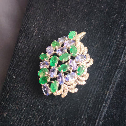 A gold-toned leaf-shaped brooch or pin embellished with green emerald-cut stones and lavender-purple oval-cut stones, resting on a textured black fabric surface.