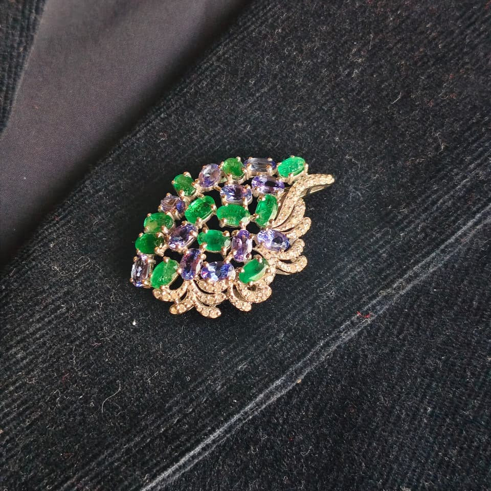 A gold-toned leaf-shaped brooch decorated with green and purple faceted gemstones, resting on a dark textured fabric background.