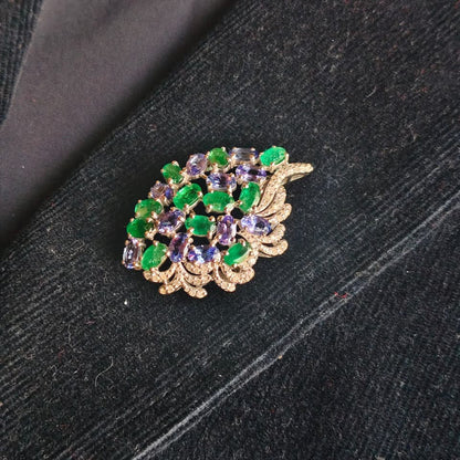 A gold-toned leaf-shaped brooch decorated with green and purple faceted gemstones, resting on a dark textured fabric background.