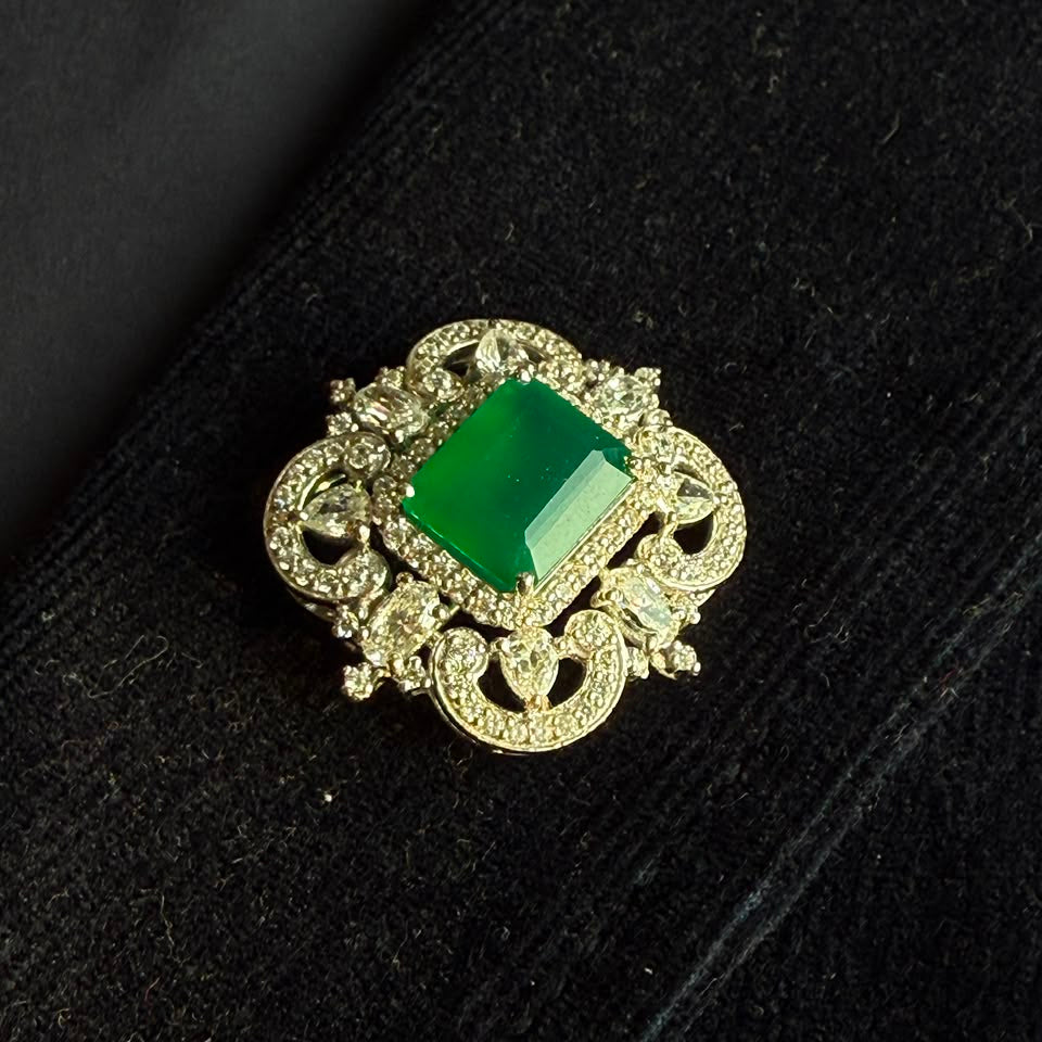 A vintage-style brooch featuring a rectangular green emerald-cut stone surrounded by ornate gold filigree and small clear stones, displayed on a black velvet surface.