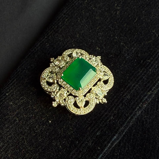 A vintage-style brooch featuring a rectangular green emerald-cut stone surrounded by ornate gold filigree and small clear stones, displayed on a black velvet surface.