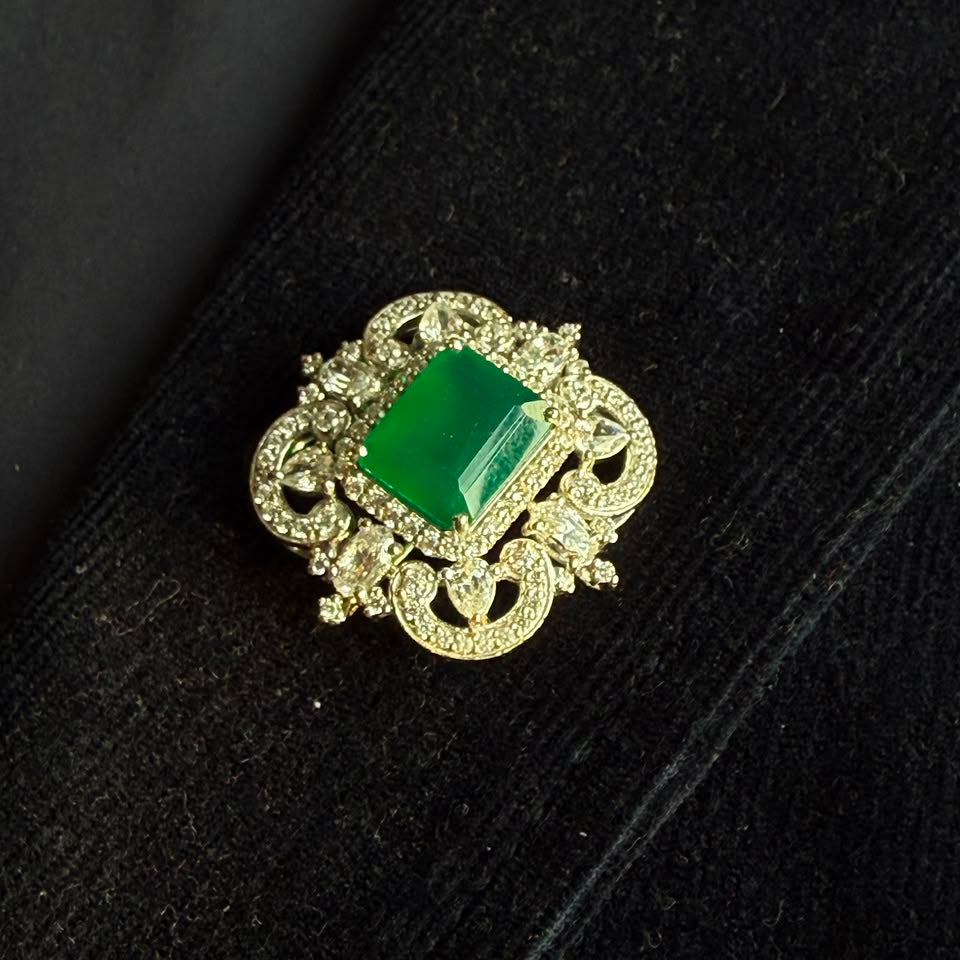 A vintage-style gold brooch featuring a prominent rectangular green gemstone surrounded by intricate filigree detailing and small clear stones, displayed on a black textured surface.