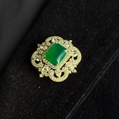 A vintage-style gold brooch featuring a prominent rectangular green gemstone surrounded by intricate filigree detailing and small clear stones, displayed on a black textured surface.