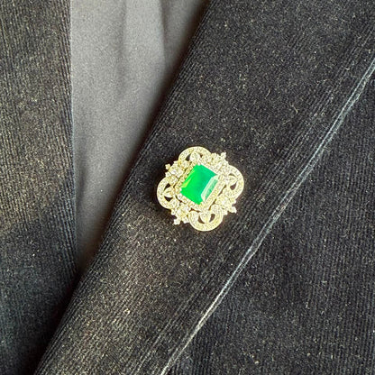 A gold-toned vintage-style brooch with a rectangular green gemstone, displayed on a dark charcoal gray textured jacket.