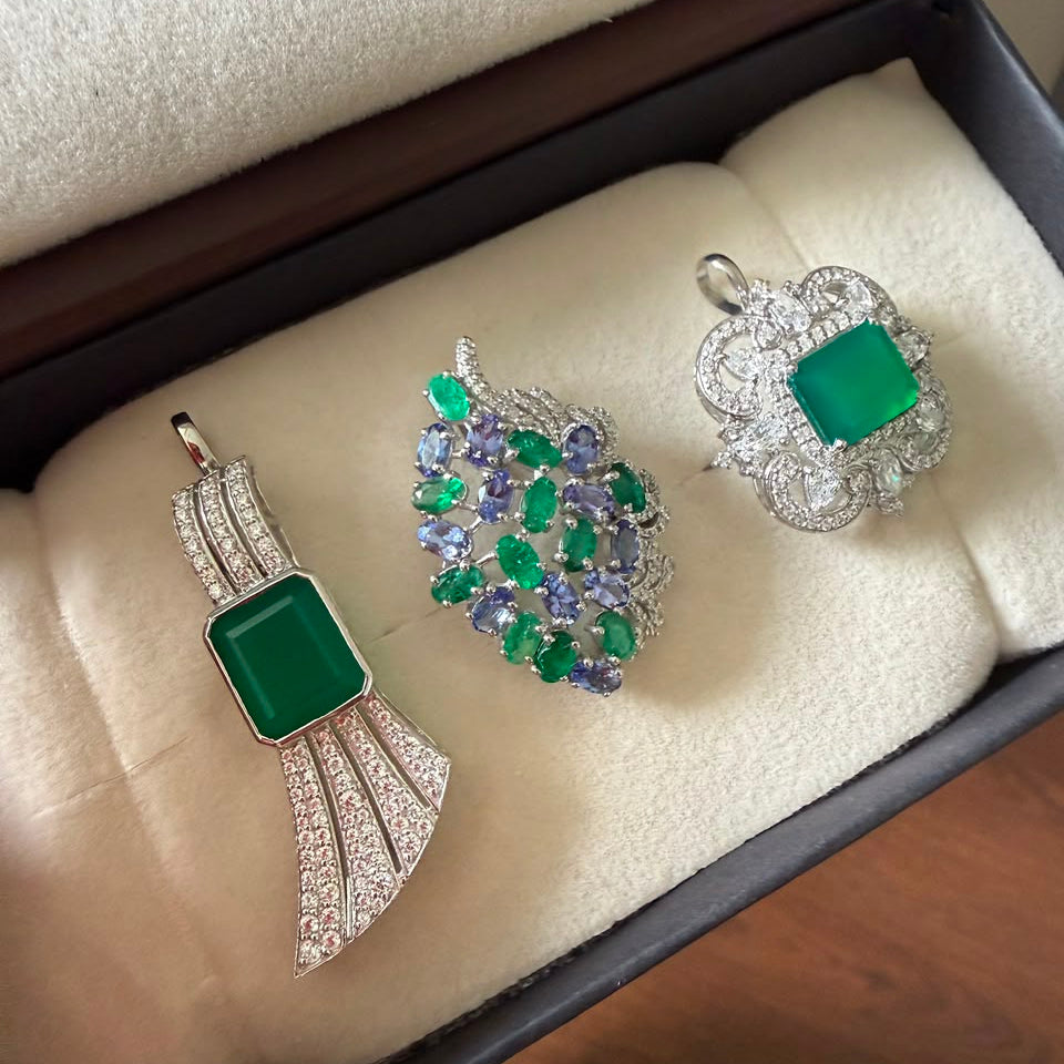 Three jewelry pieces displayed in a jewelry box: a silver pendant with a green rectangular stone, a silver and green leaf-shaped brooch with purple and green stones, and a silver ring with a green rectangular stone and clear accents.