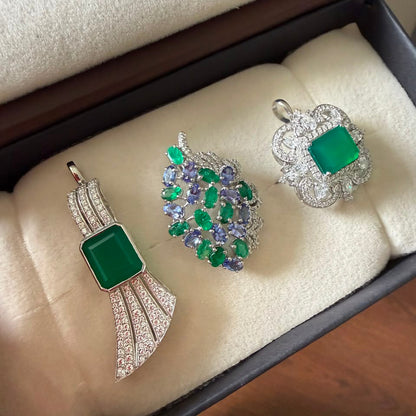 Three jewelry pieces displayed in a jewelry box: a silver pendant with a green rectangular stone, a silver and green leaf-shaped brooch with purple and green stones, and a silver ring with a green rectangular stone and clear accents.