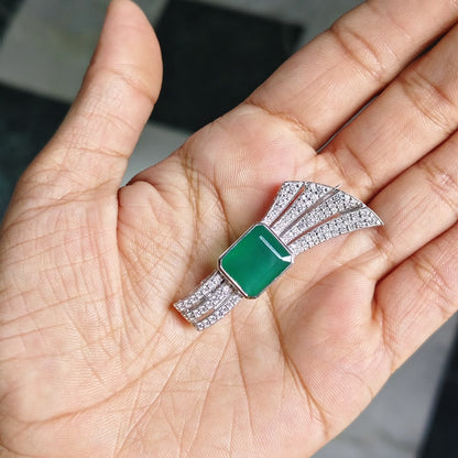 A close-up of a hand holding a silver-toned fan-shaped brooch with a rectangular green jade-like center stone and small clear stones along the edges, against a black-and-white checkered floor background.
