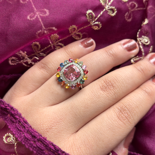 A close-up of a hand wearing a colorful ring with a large pink cushion-cut center stone surrounded by multicolored gemstones, set against a rich magenta embroidered fabric background.