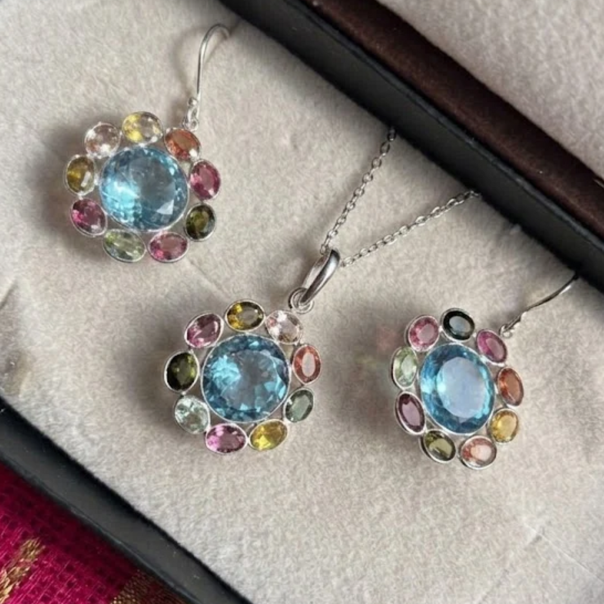 A jewelry set featuring a necklace and two matching drop earrings with silver-toned metal and a central round light blue stone surrounded by small multicolored accent stones, displayed on a beige jewelry box insert.
