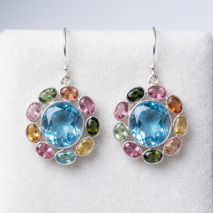 A pair of silver dangle earrings with large round light blue center stones surrounded by a halo of small multicolored oval gemstones, displayed on a white textured surface.